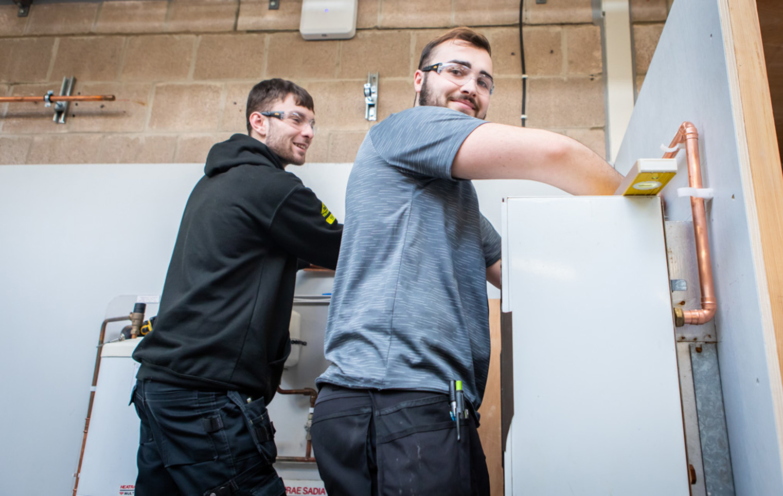 Two plumbing students smiling while collaboratively working on installing a plumbing fixture, engaged in hands-on training.