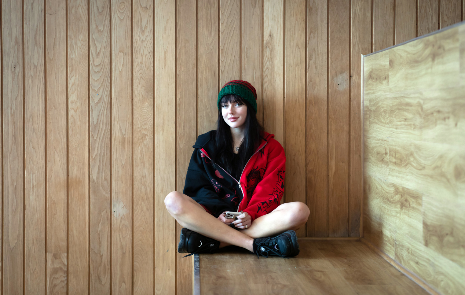 Student sitting cross-legged on a wooden bench against a wood-panelled wall at Glasgow Kelvin College’s Springburn Campus, wearing a red and black jacket and green hat.
