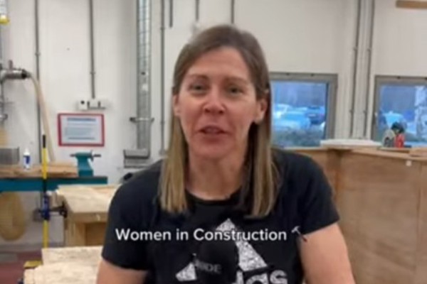 A female lecturer in a black Adidas t-shirt speaking in a joinery workshop at Glasgow Kelvin College, introducing the 'Women in Construction' course, with workbenches and tools visible in the background