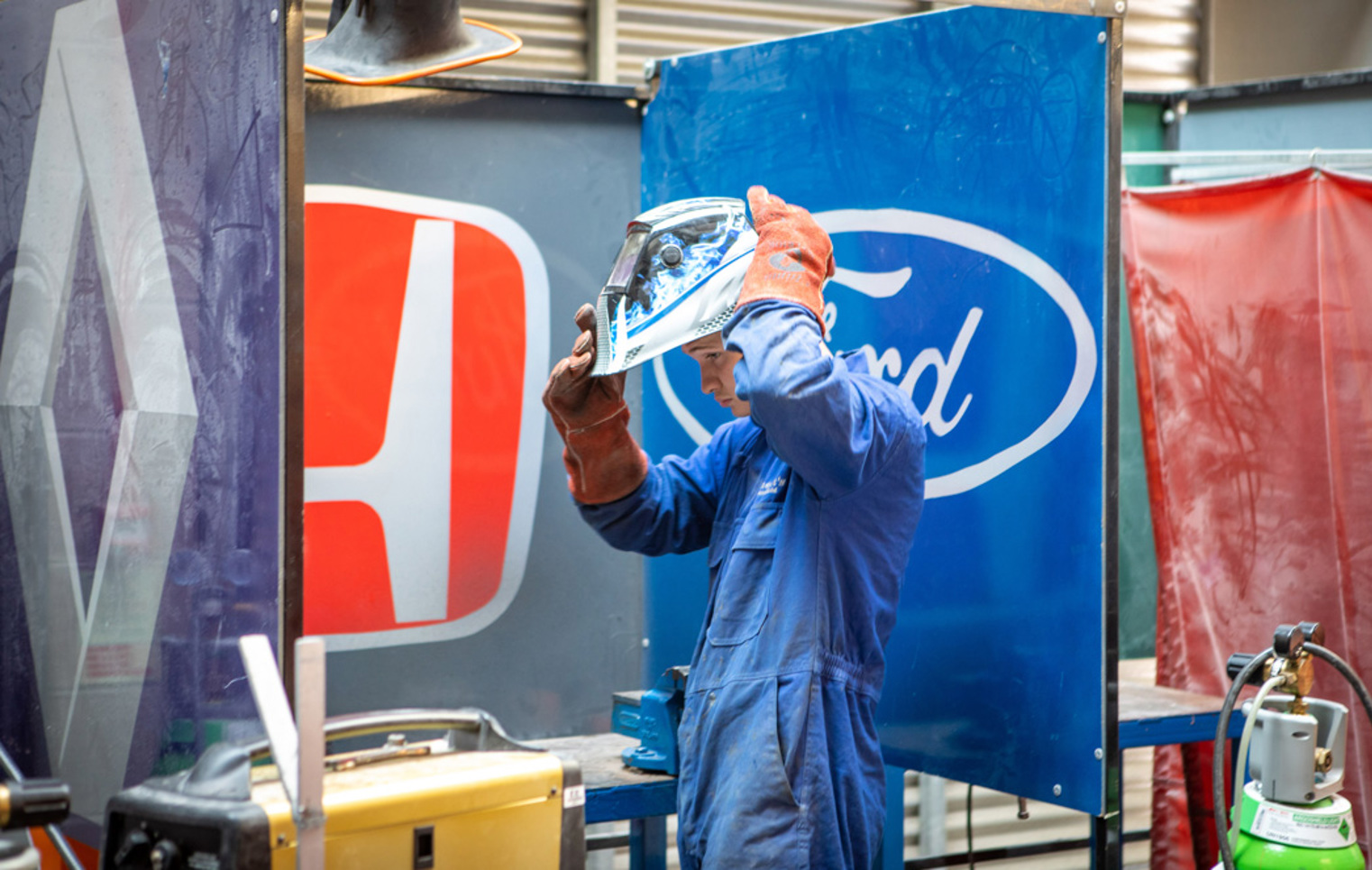 Student adjusting welding helmet beside car manufacturer screens in the automotive workshop.