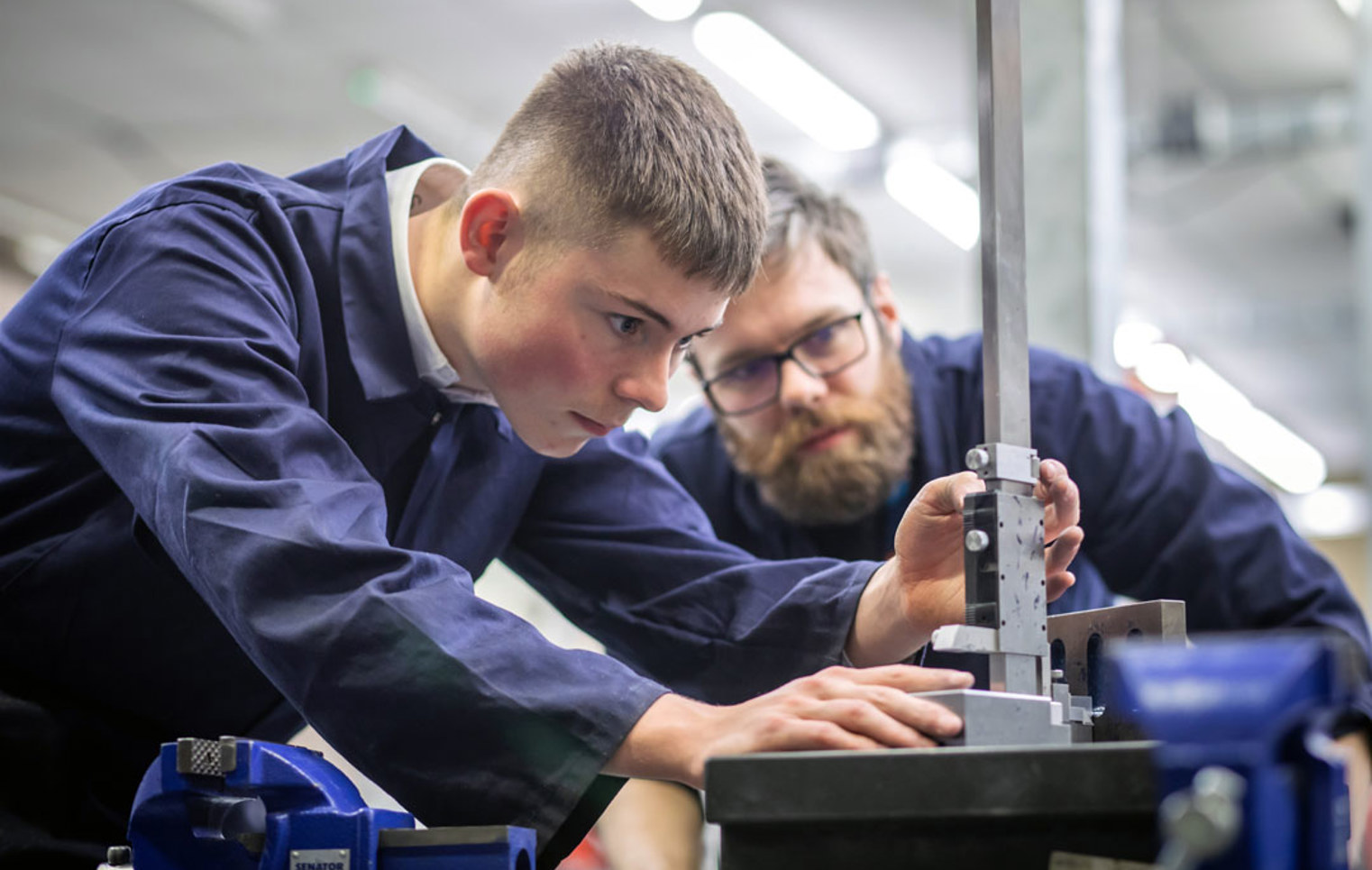 A lecturer and a student in navy-blue overalls working together in a mechanical engineering workshop. The lecturer is demonstrating how to use a tool on a metal component held in a vise.
