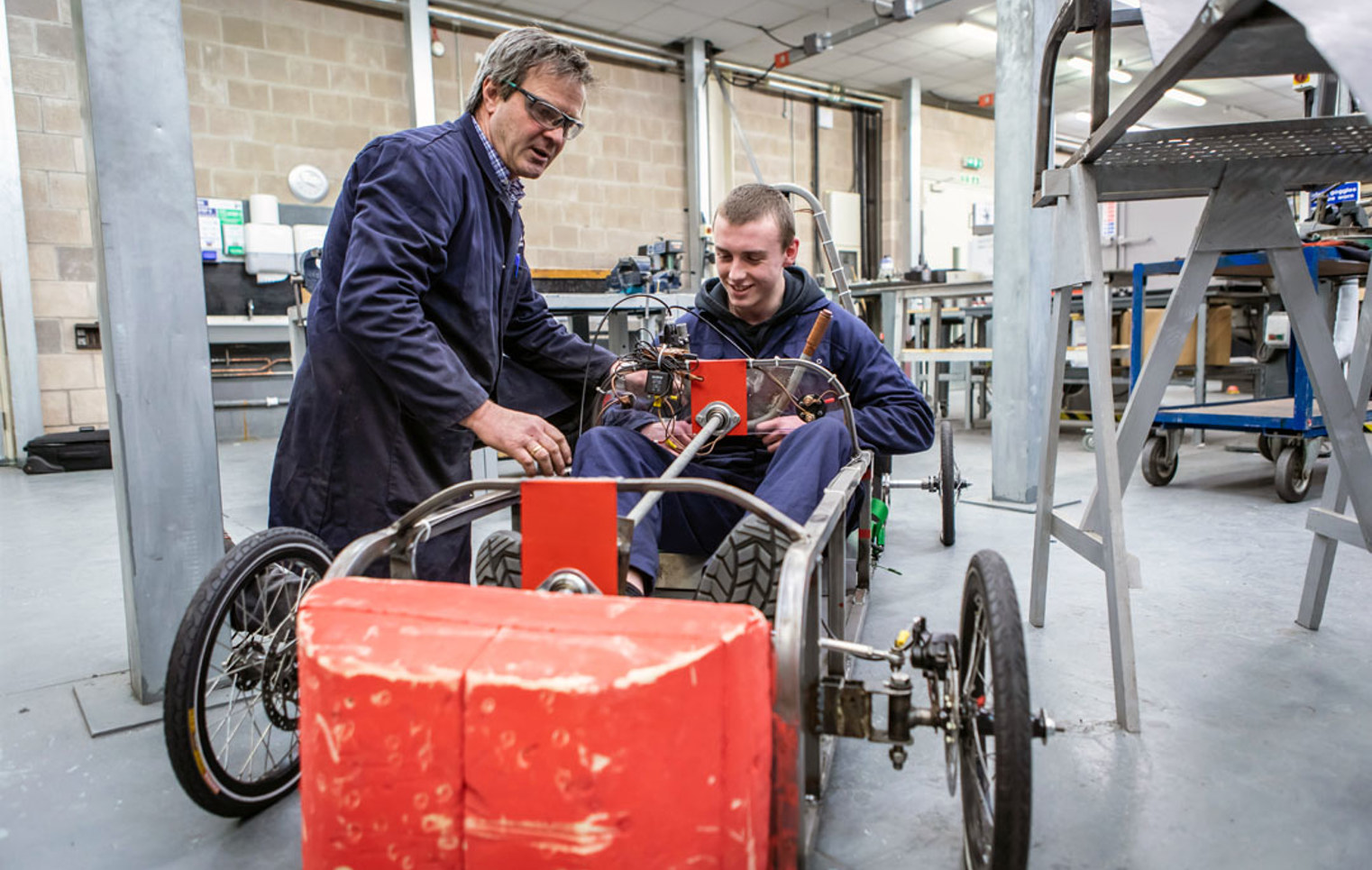 A lecturer assisting a student seated in a custom-built electric car, adjusting the wiring and components during a hands-on engineering project.