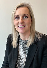 Joanna Campbell smiling, wearing a black blazer and patterned top, seated against a plain light background.