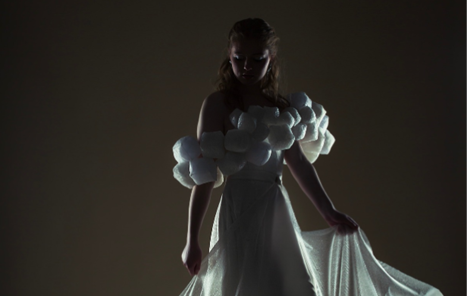 Model in a dramatic, low-lit setting wearing an off-the-shoulder white gown with textured puff sleeves and a flowing sheer train, softly illuminated from behind.