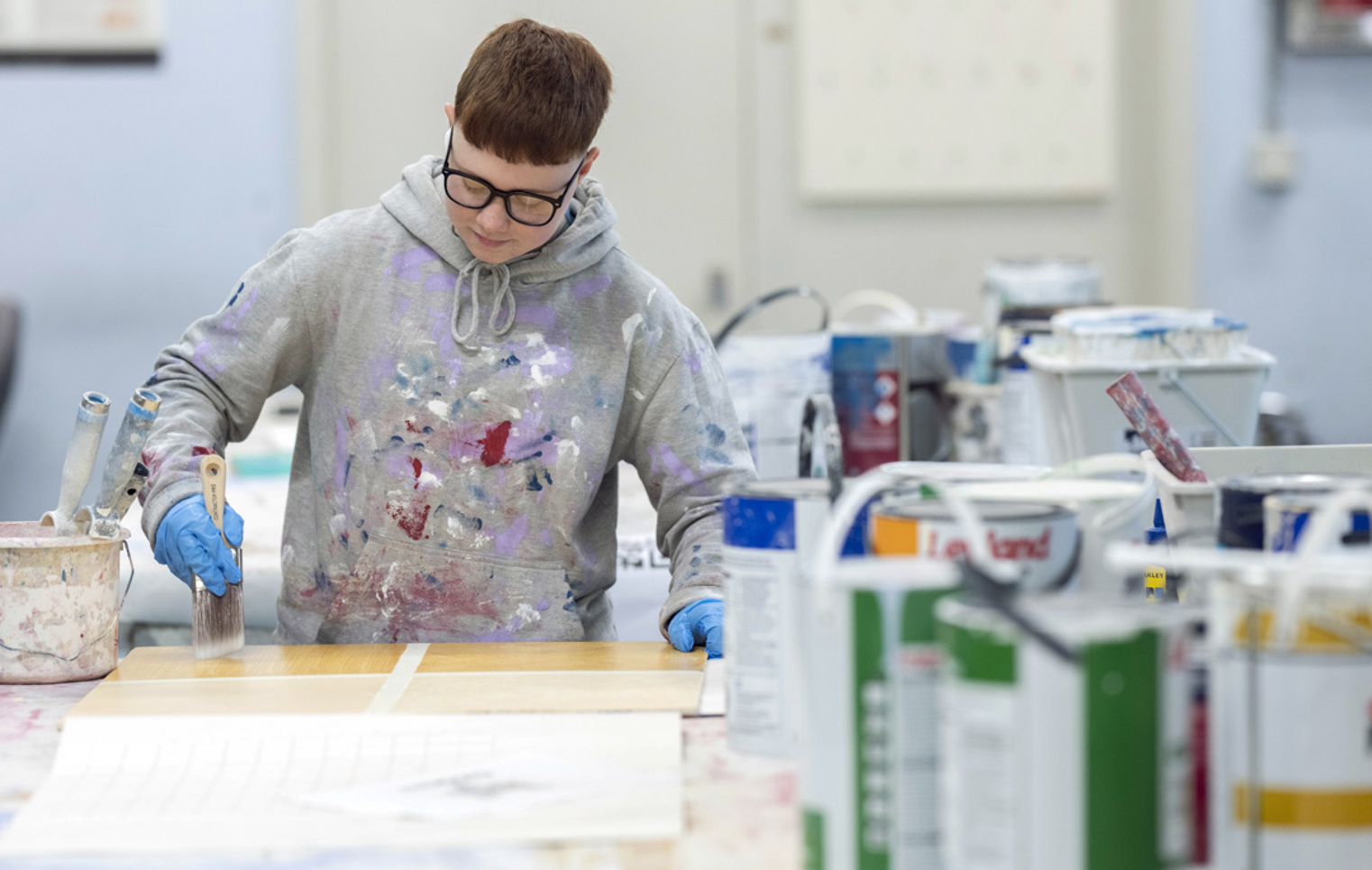 Focused student in paint-splattered hoodie painting on a wooden board