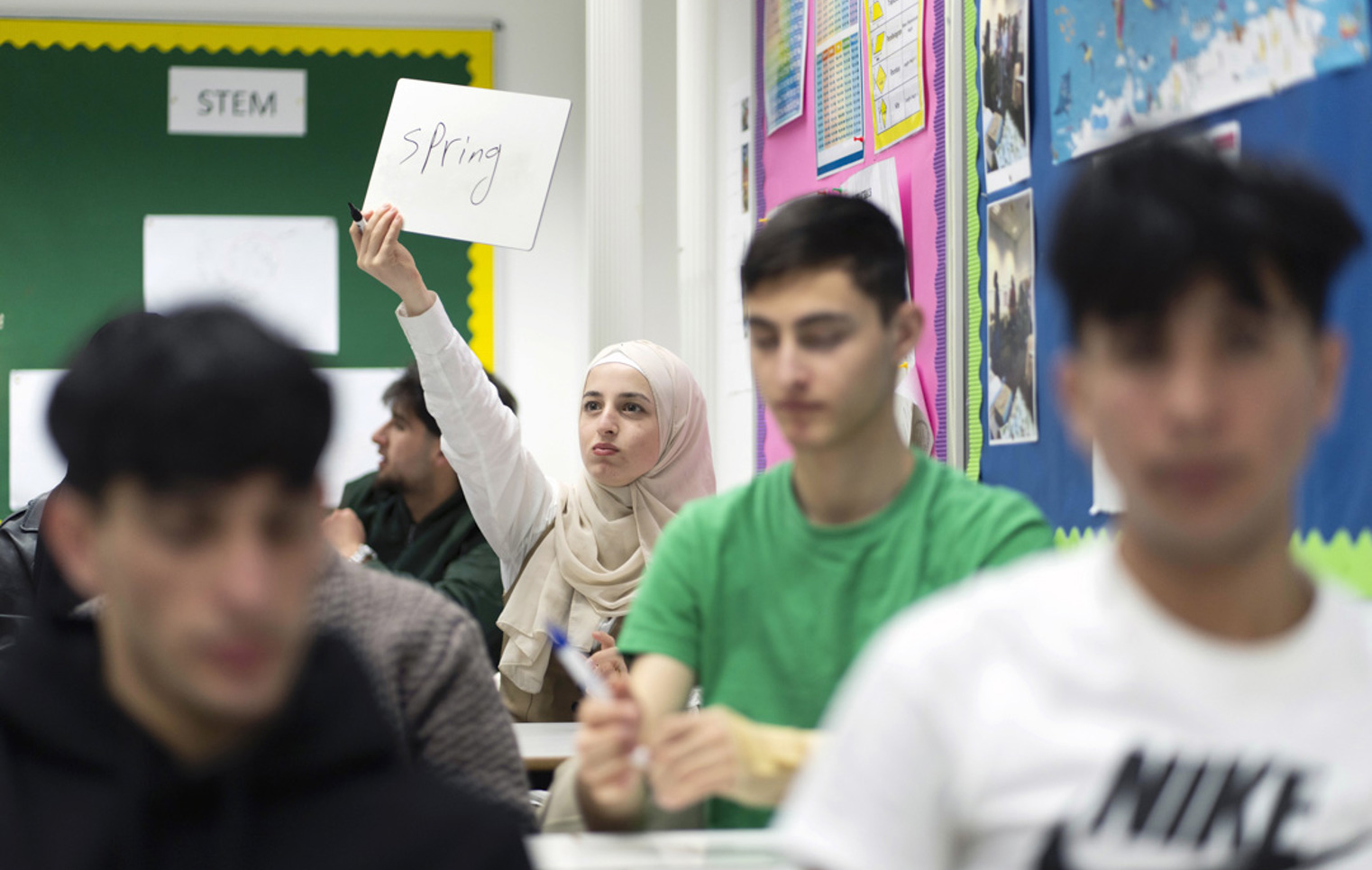 A female student in a beige hijab holds up a small whiteboard with the word "Spring" written on it, participating in a classroom activity. Other students are in the foreground, slightly out of focus.