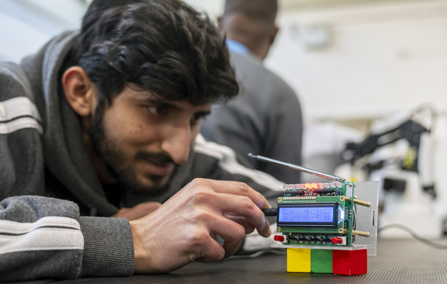 Student closely examining a custom-built radio circuit board with LED indicators and an antenna in an electronics lab