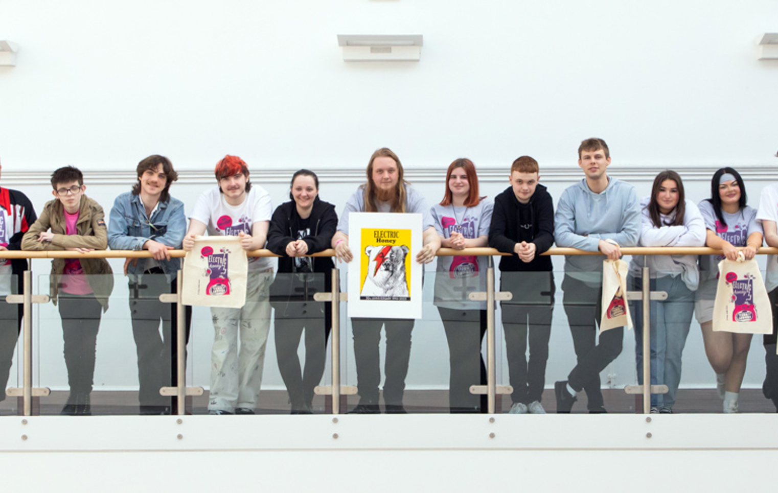Group of music business students and a lecturer lined up along a balcony, some holding Electric Honey tote bags and a framed anniversary poster.
