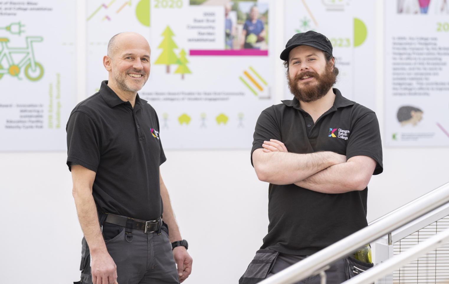 Two male staff members from the Glasgow Kelvin College Estates Team standing indoors, smiling at the camera in front of sustainability-themed posters.