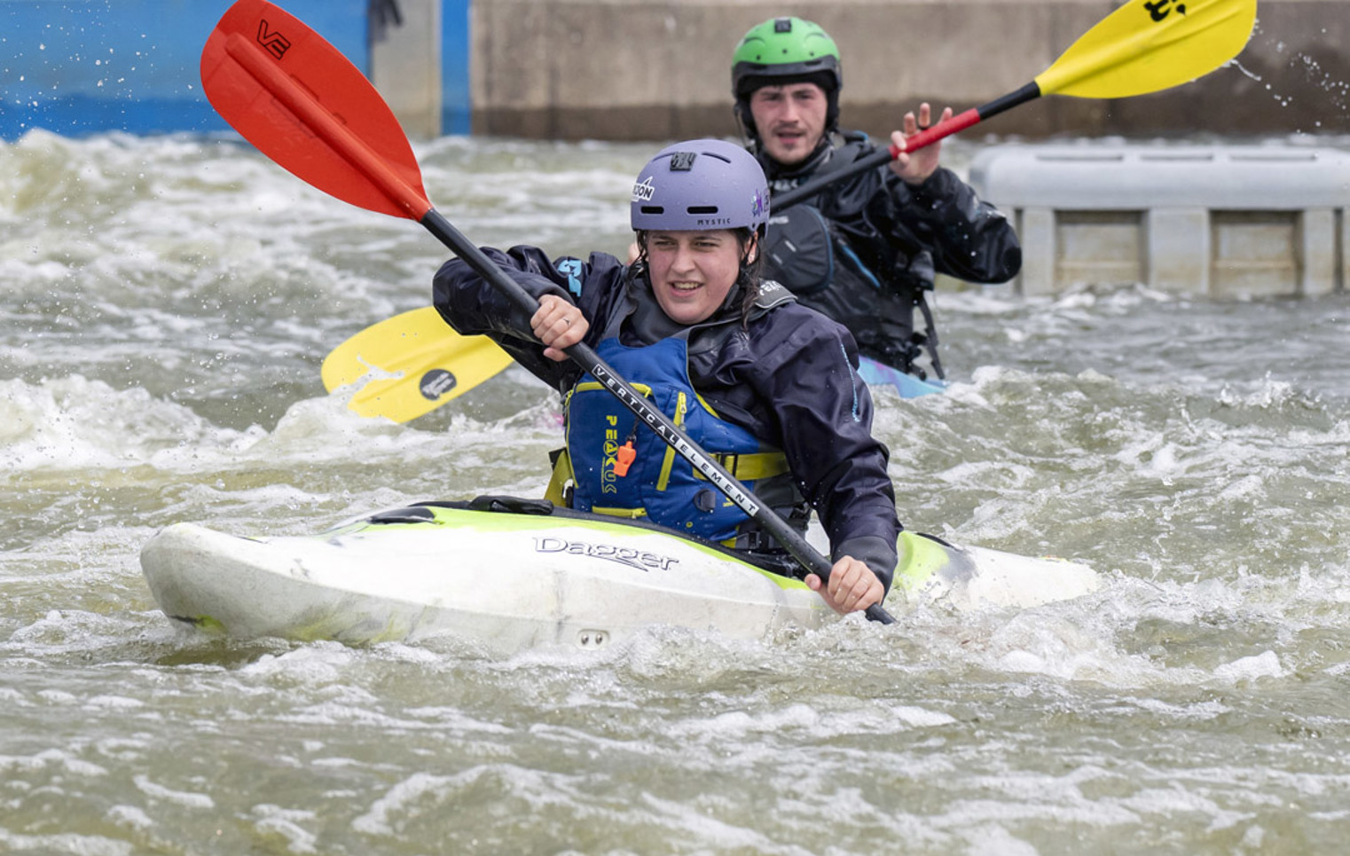 Two people kayaking in whitewater rapids, wearing helmets and waterproof gear during an outdoor activity session.