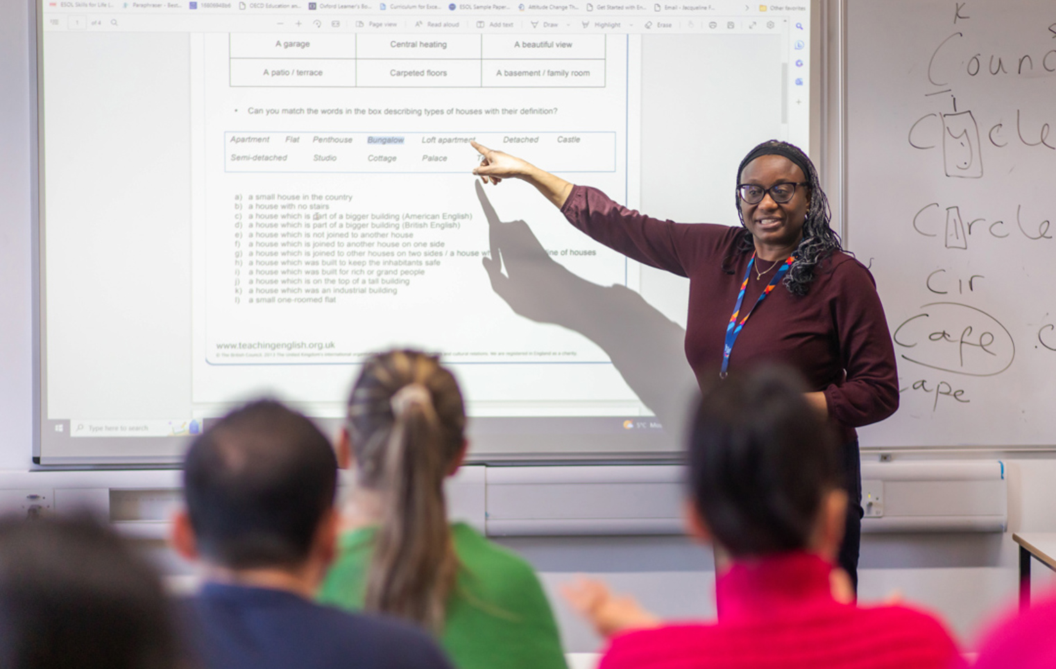 A female lecturer wearing glasses and a lanyard points to a projected lesson on a whiteboard while teaching a group of students.