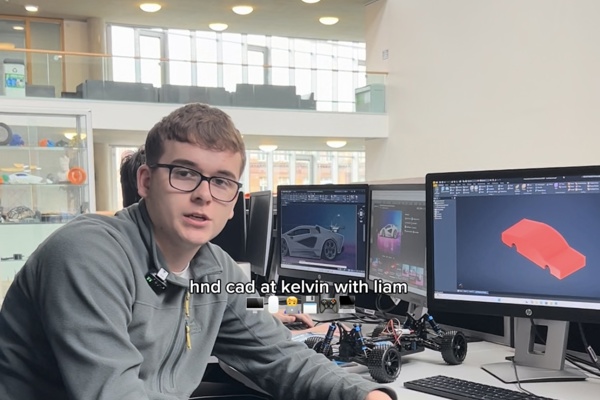 HND CAD student Liam sitting at a computer desk in Glasgow Kelvin College, with 3D car models displayed on dual monitors and a small vehicle prototype on the table.