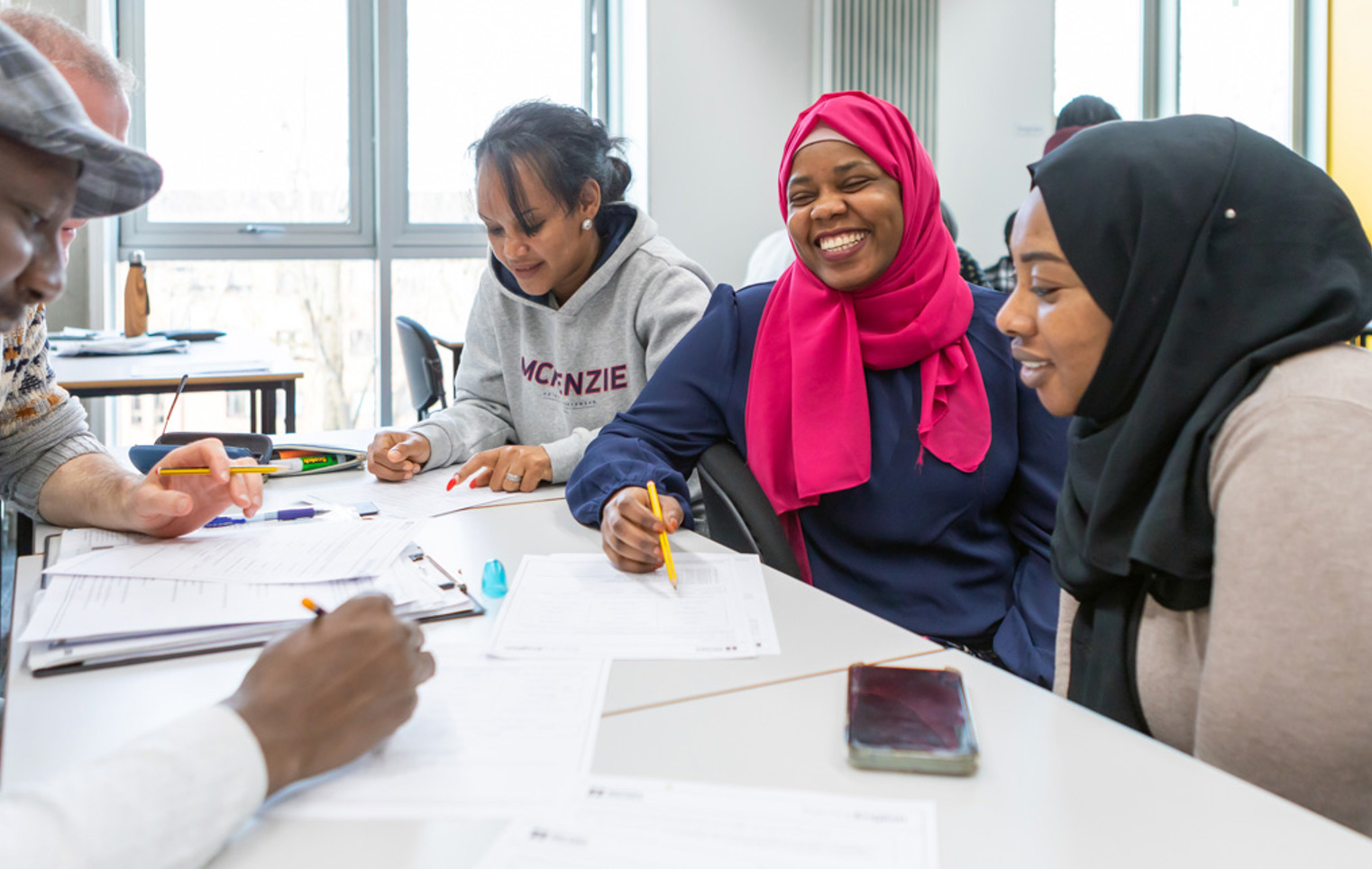 A diverse group of students, including a woman in a pink hijab, laughs and collaborates while working on an assignment in a classroom.
