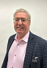Martin Booth, a man wearing glasses, a pink shirt, and a blue check blazer, smiling against a light background.
