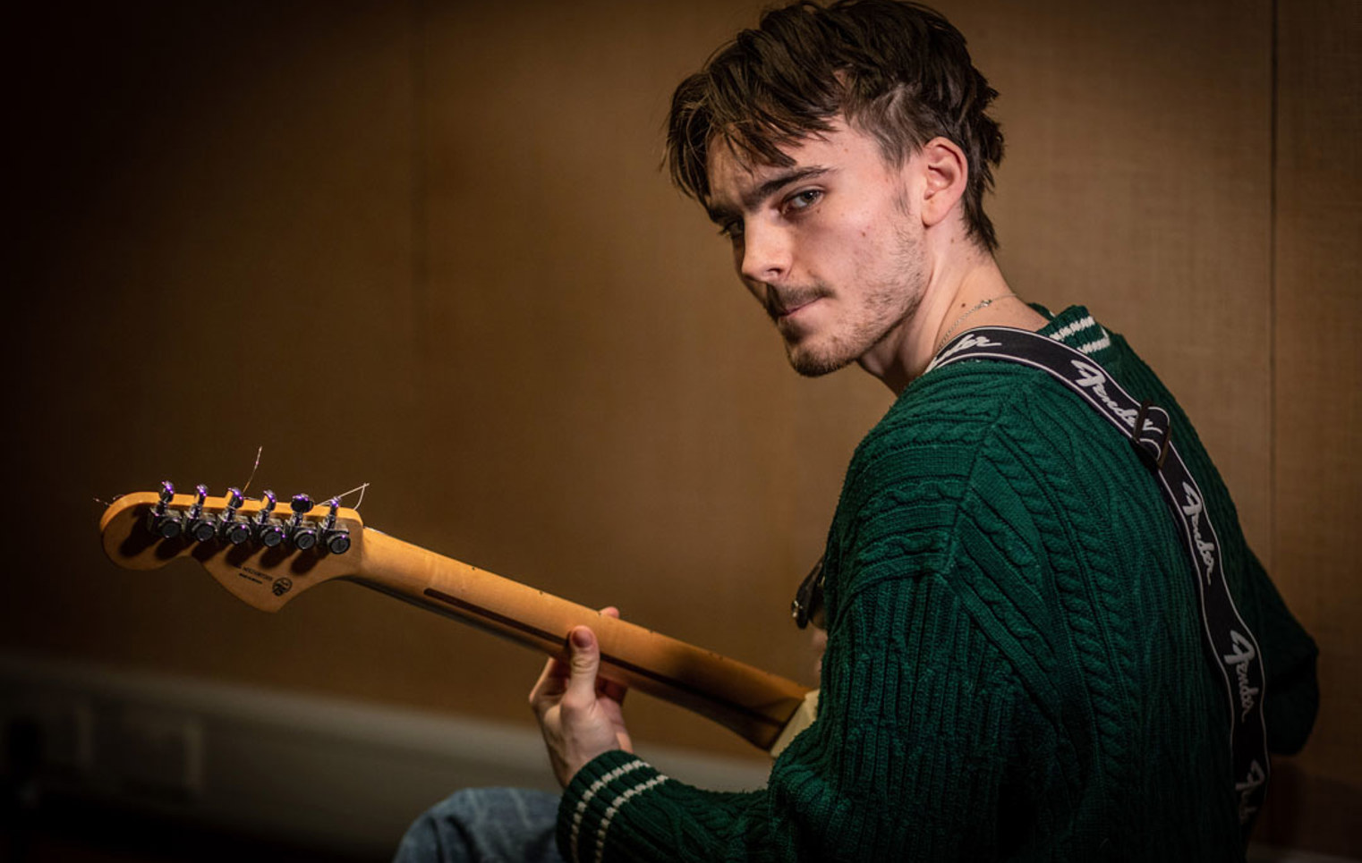 A young guitarist wearing a green sweater, holding a Fender guitar, and looking towards the camera in a dimly lit practice room.