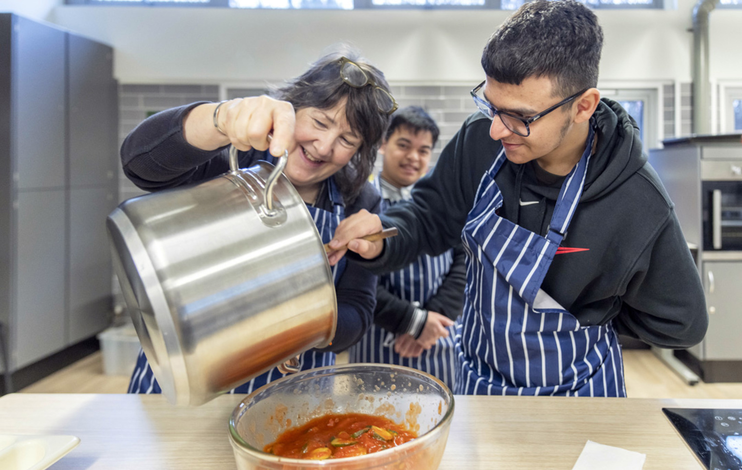 Woman pouring sauce from a large pot into a bowl while smiling and working with two young men in striped aprons.