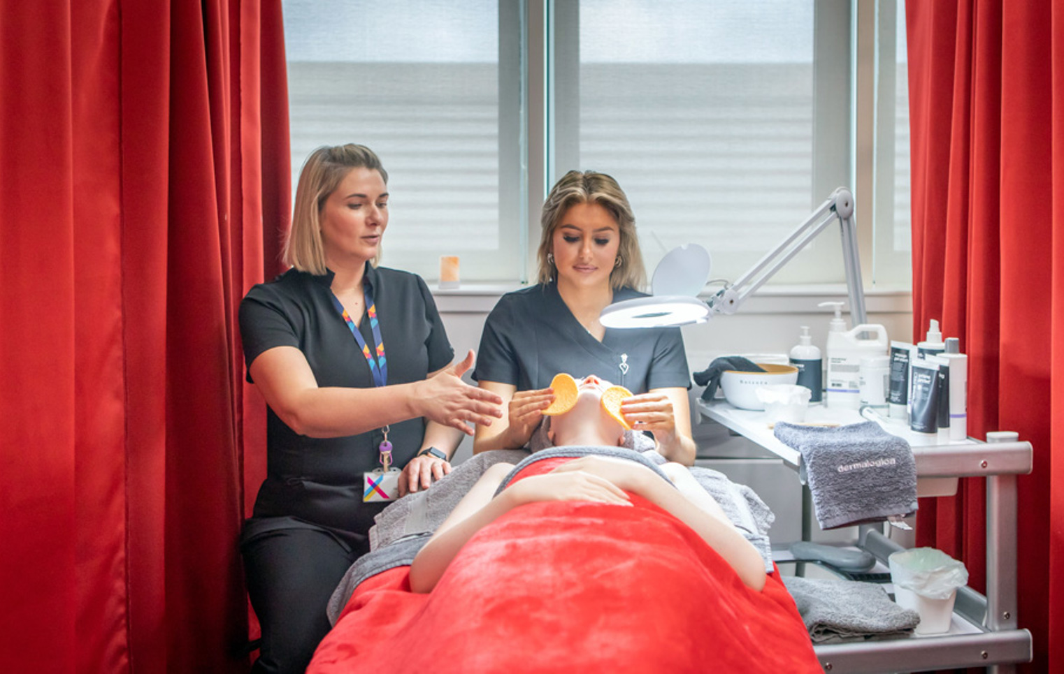 A lecturer demonstrates a facial massage technique to a beauty therapy student who is performing a treatment on a mannequin in a spa-like environment.