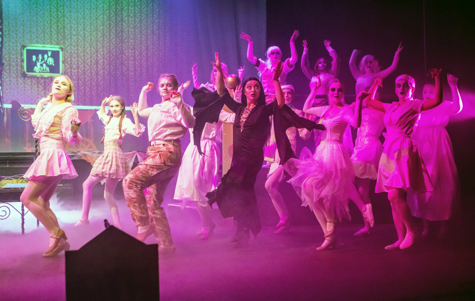 Musical theatre students performing a high-energy ensemble dance on stage, with raised arms, coordinated movement, and purple lighting.