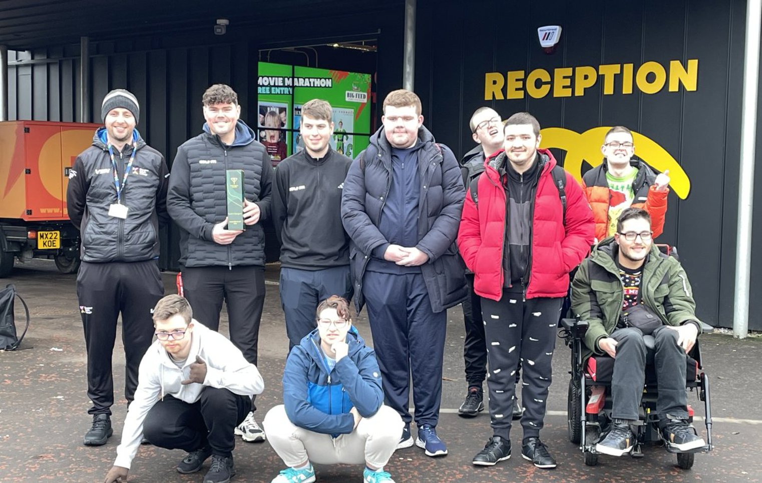 Group of Glasgow Kelvin College students and staff smiling outside the Golf It! reception, some crouching and one in a wheelchair, after a visit.