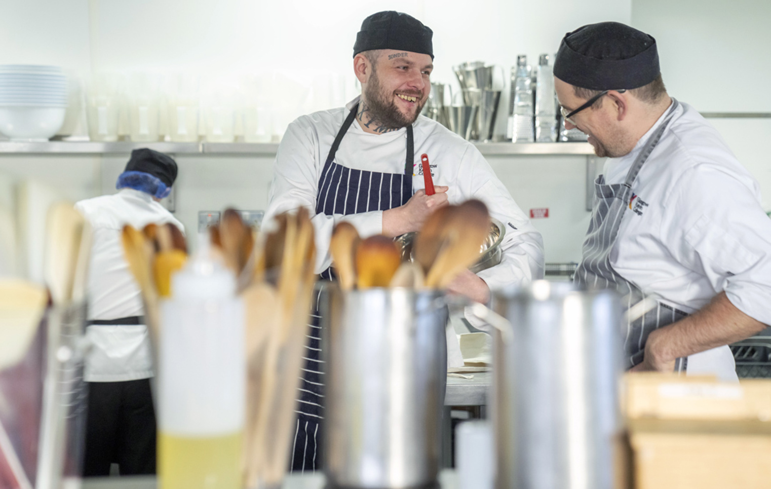 A chef with tattoos and a striped apron smiles while chatting with an instructor in a busy kitchen, with cooking utensils and supplies in the foreground.