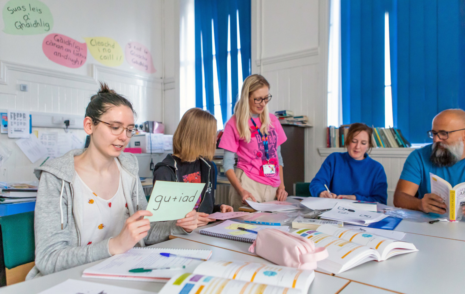 Gaelic lecturer supporting students in a language learning activity, with students holding vocabulary cards and engaging in conversation.