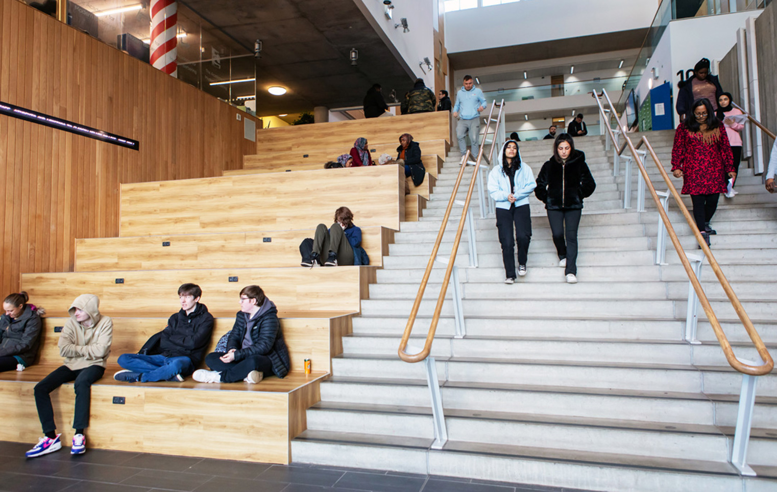 Students sitting and walking on the main staircase and tiered wooden seating area at Glasgow Kelvin College’s Springburn Campus.