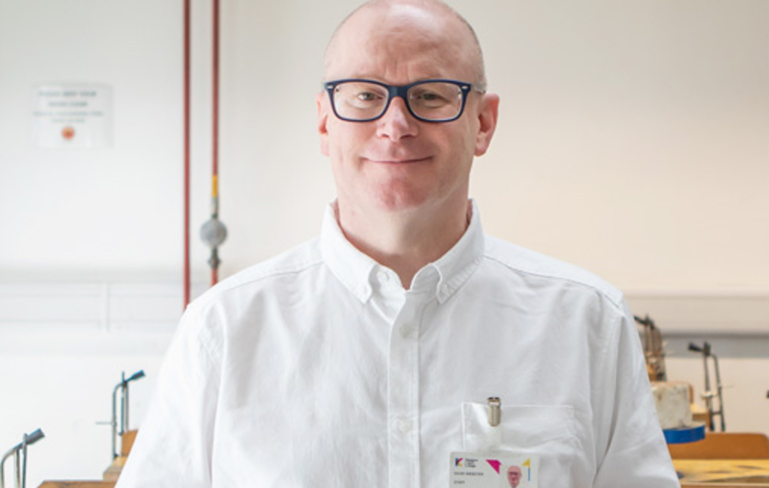 Lecturer David Webster stands in a jewellery workshop at Glasgow Kelvin College, smiling and holding a jeweller’s saw, with tools and benches in the background.