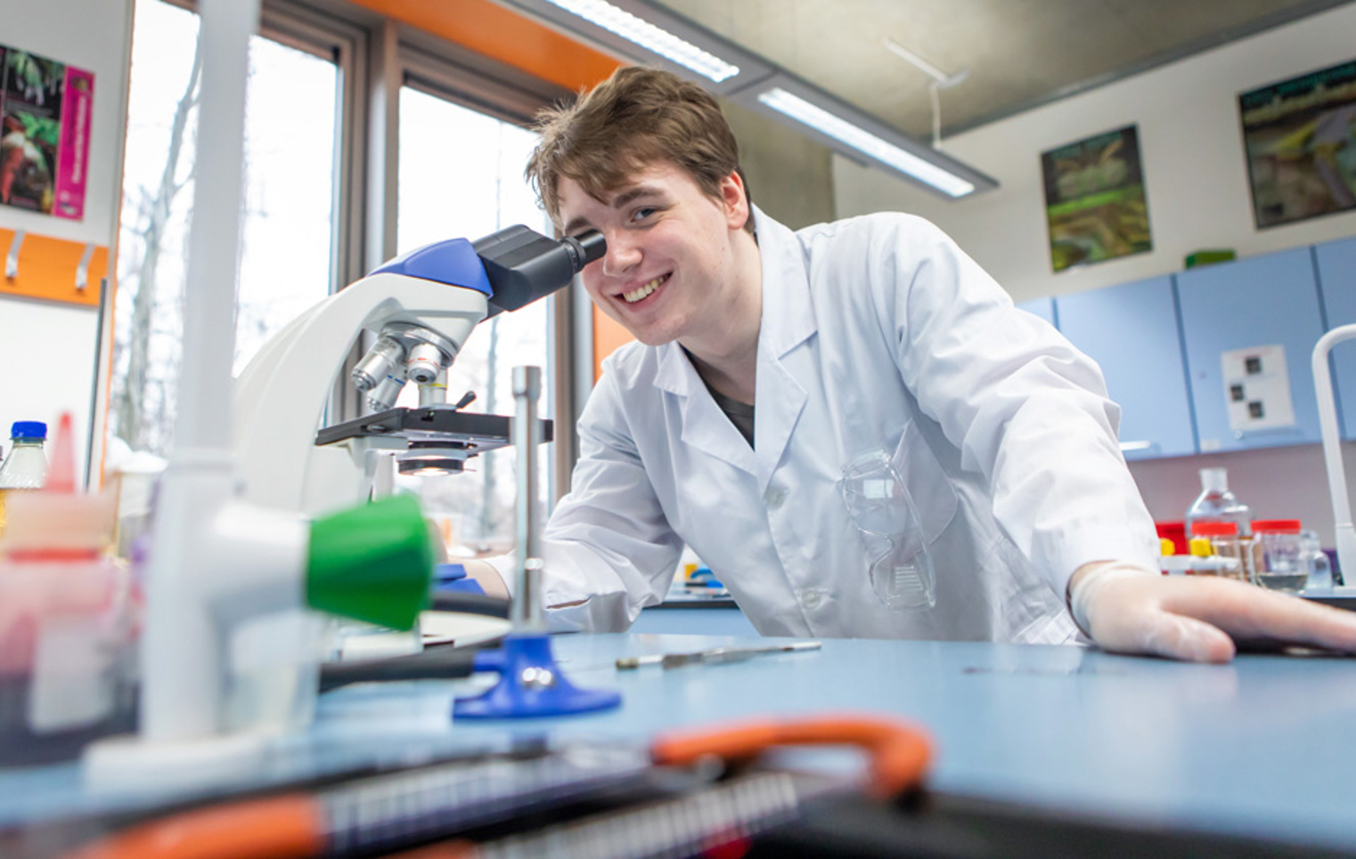 Student using a microscope in a science classroom filled with lab equipment and colourful bottles.