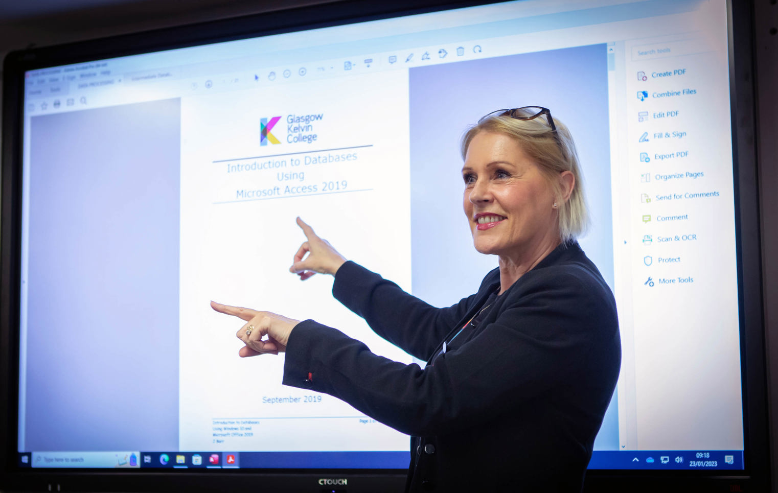 Lecturer in a black blazer presenting a lesson on Microsoft Access, pointing at a large interactive screen in a computer lab.