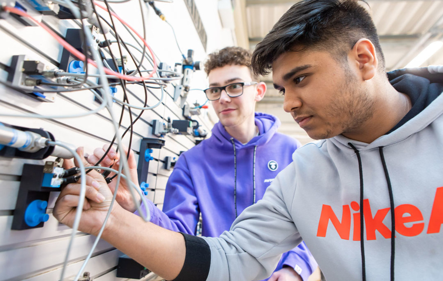 Two students working together on a pneumatics system, connecting wires and tubes to an industrial training panel.