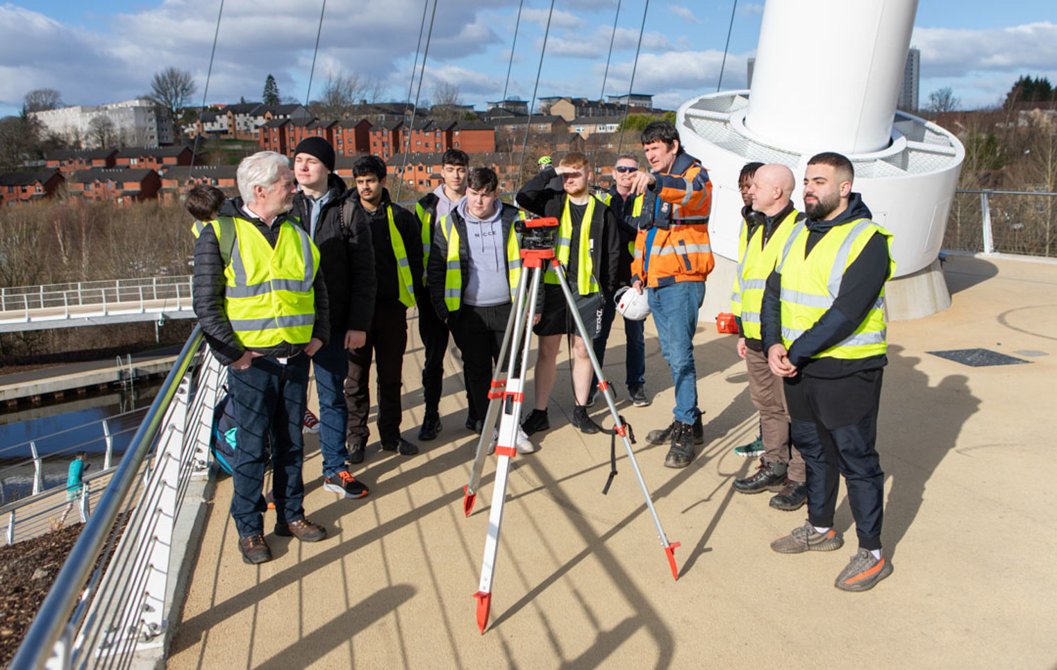 A group of students and lecturers in high-visibility vests gathered around a surveying instrument on the Stockingfield Bridge, listening to an instructor providing guidance.