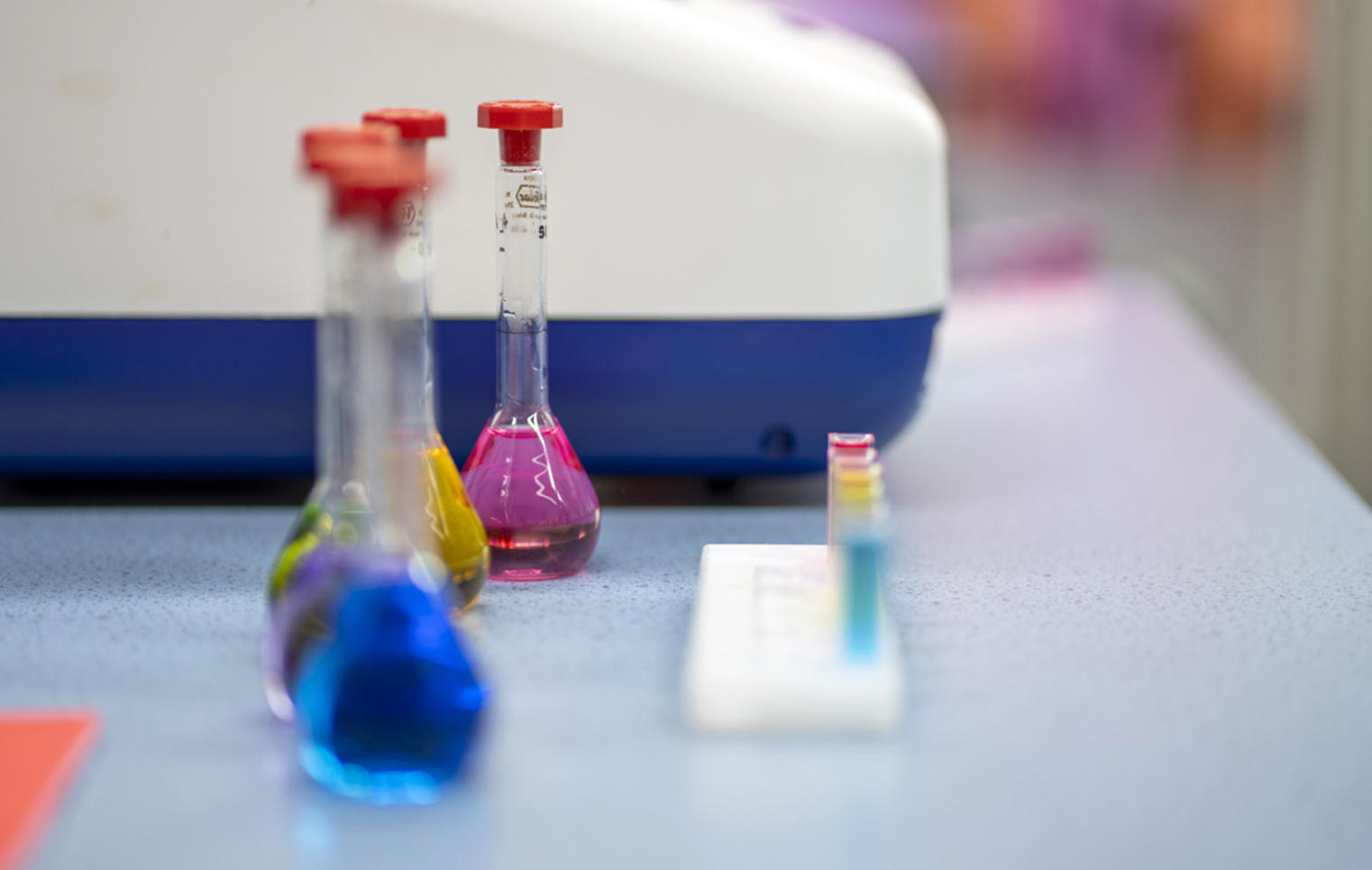 Three glass flasks filled with bright blue, yellow, and pink liquids sit on a lab bench, with a row of colourful test tubes and a science machine in the background—ready for a splash of science fun and discovery.