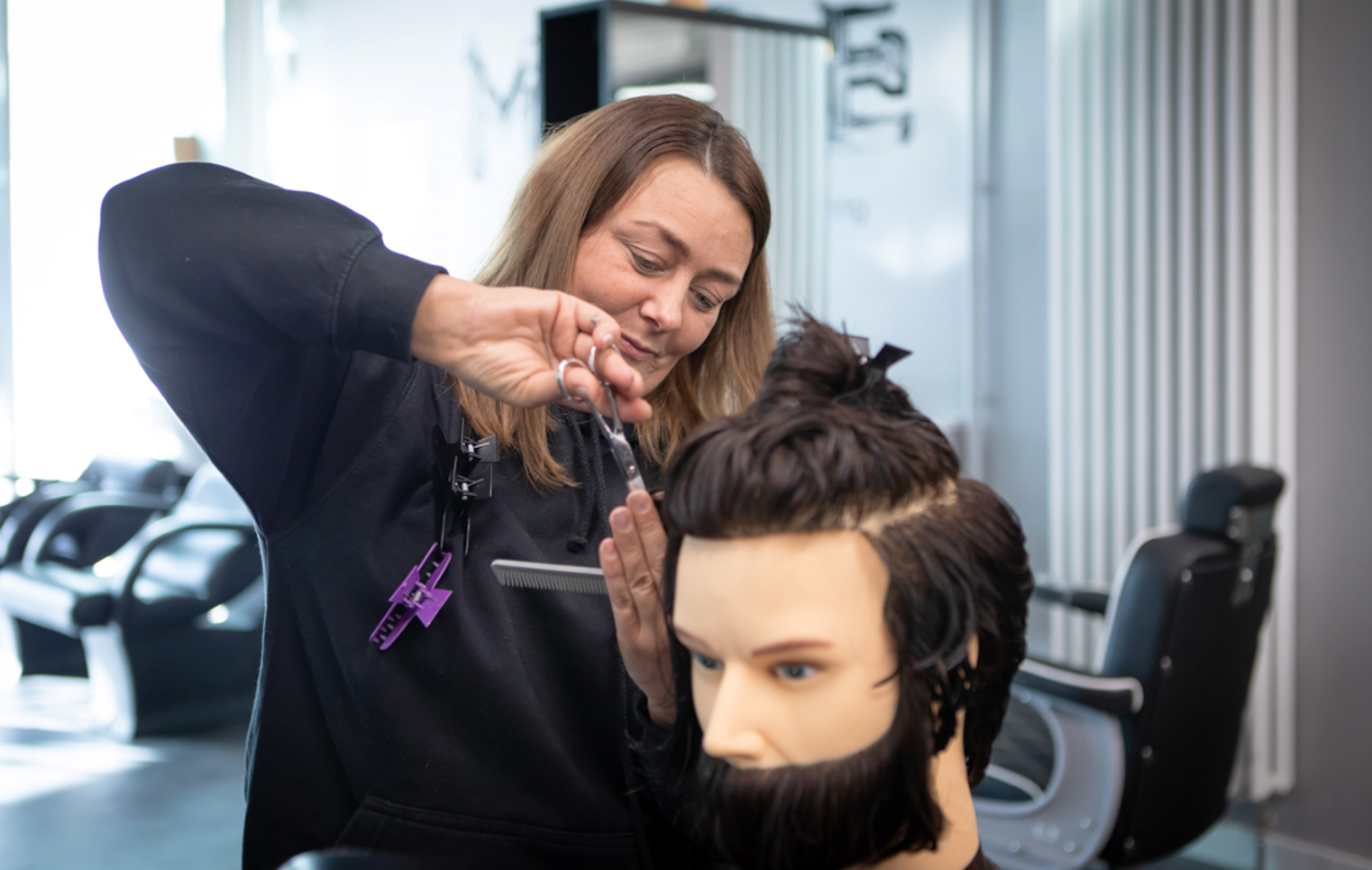A female barbering student in a black hoodie carefully cutting the hair on a mannequin head inside a training facility.