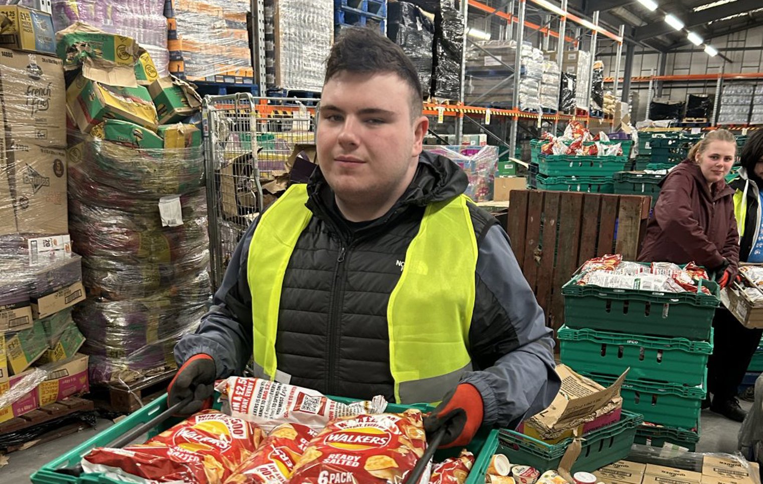 Young man wearing a high-visibility vest holding a crate of packaged crisps while working in a warehouse environment.
