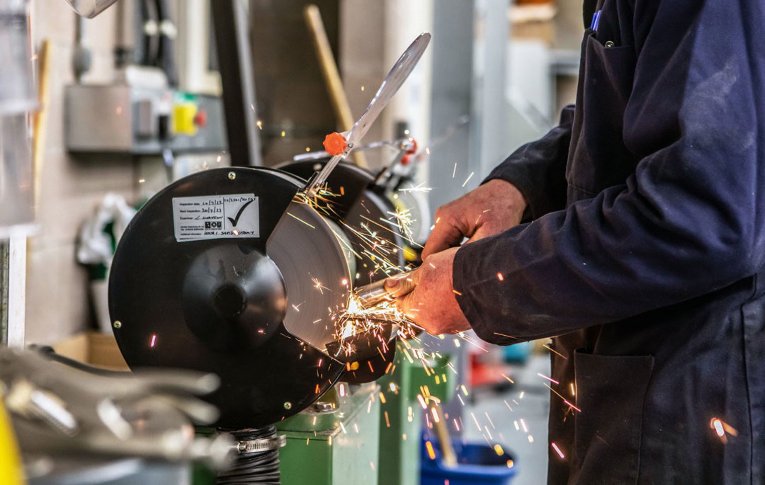 Close-up of a grinding wheel in action, with sparks flying as a lecturer in a navy-blue coat sharpens a metal component.