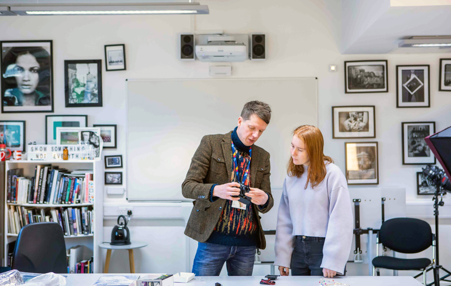 Lecturer demonstrating camera functions to a student in a photography classroom filled with artwork.