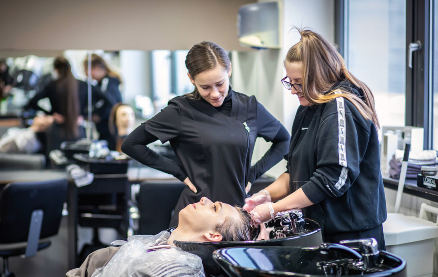 Two hairstyling students washing a client's hair at a salon sink.