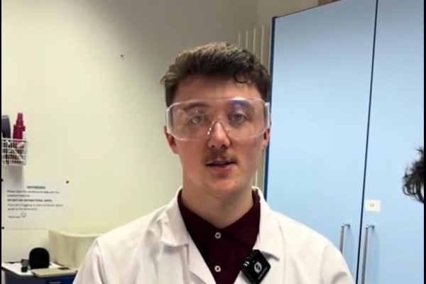 Science student Lewis wearing safety goggles and gloves while demonstrating lab equipment in a college laboratory.