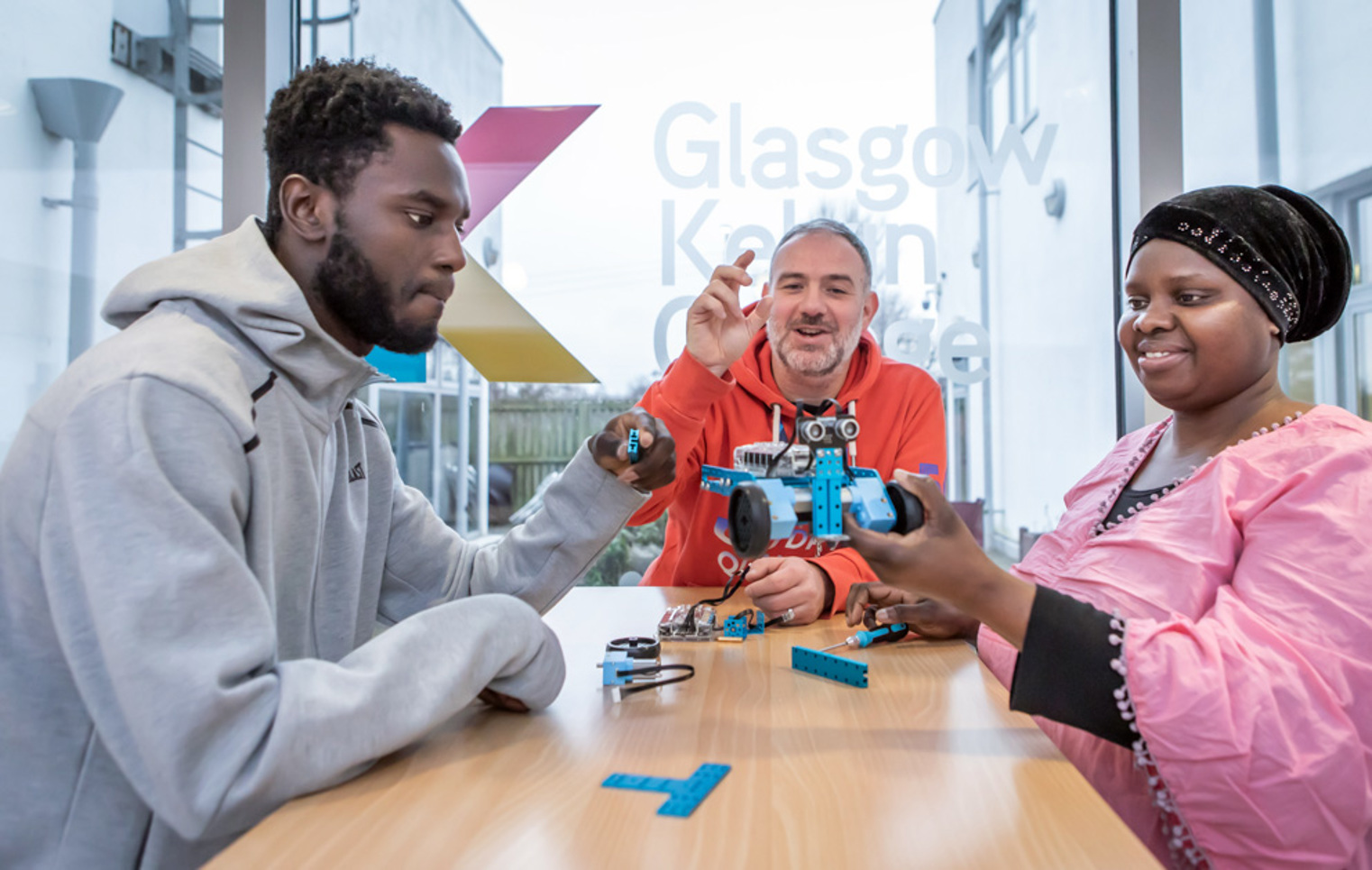 Lecturer in an orange hoodie helping two students troubleshoot and assemble a blue robotic vehicle in a classroom.