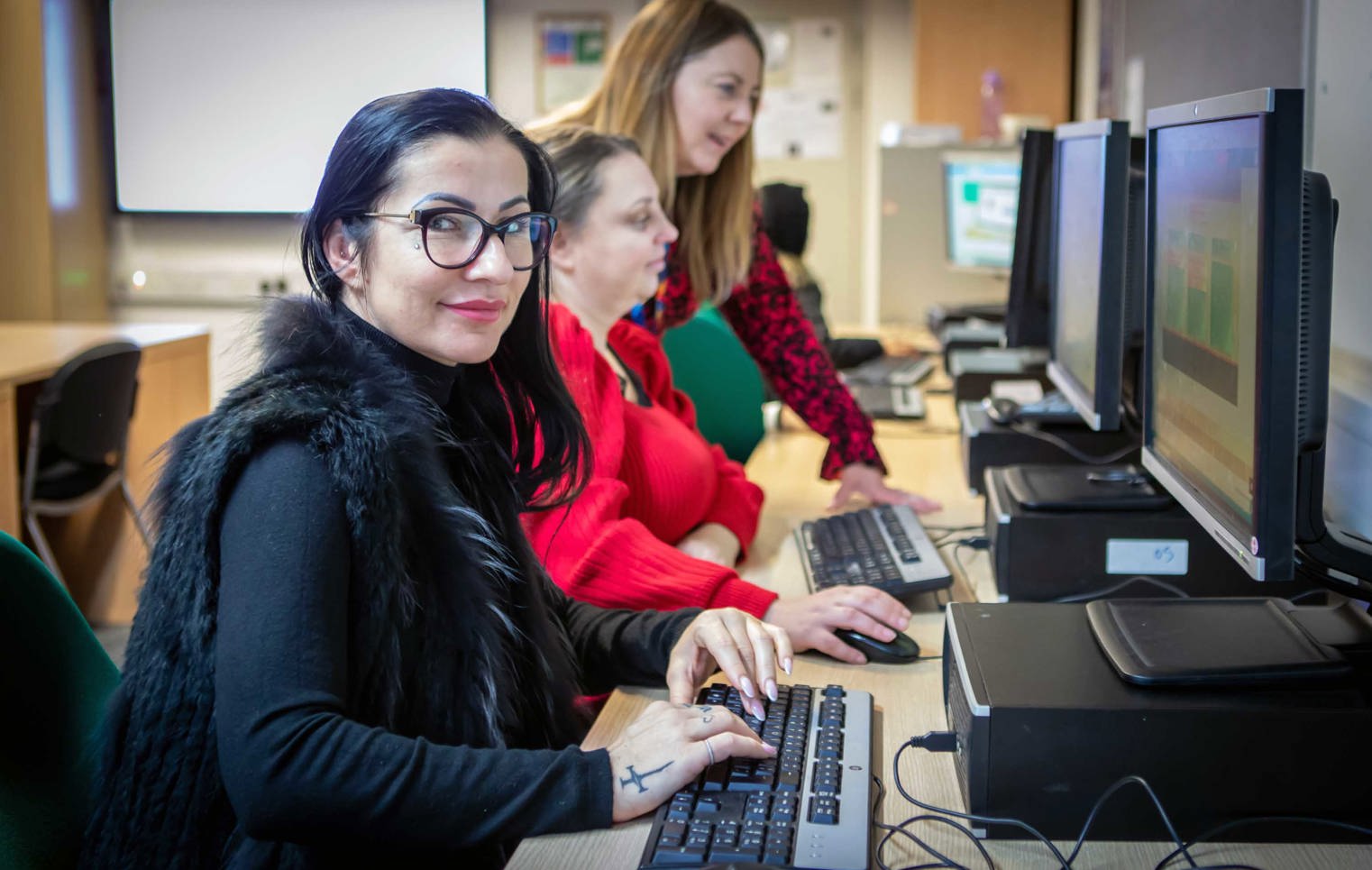 Student in a black vest working at a computer while a lecturer provides guidance and feedback in a classroom setting.