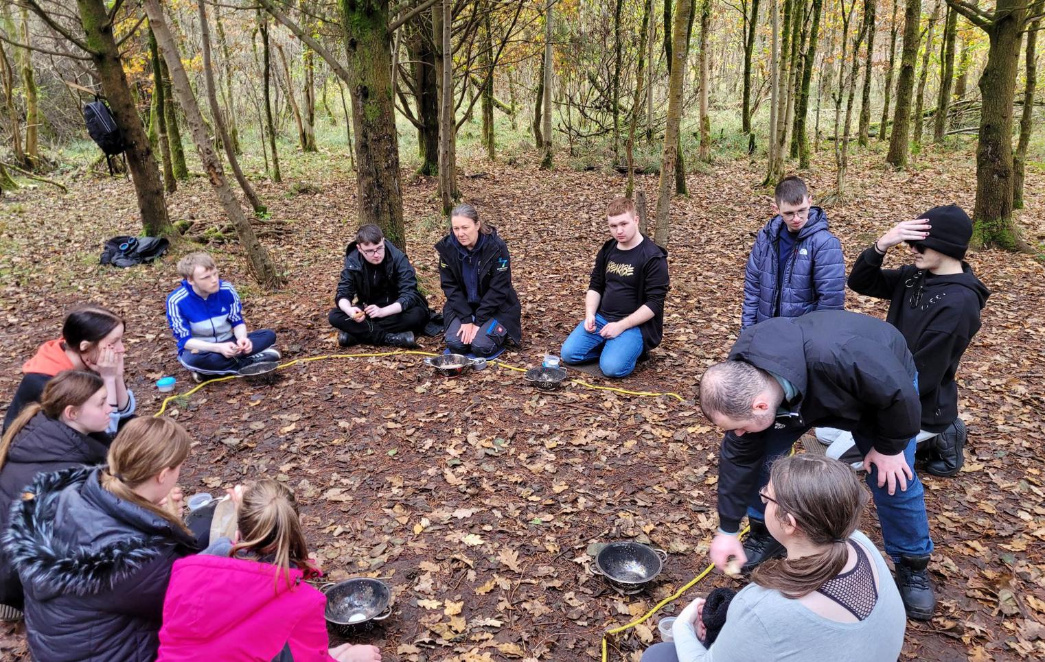 Group of young people sitting in a circle on the forest floor, engaged in a team-building or outdoor activity.