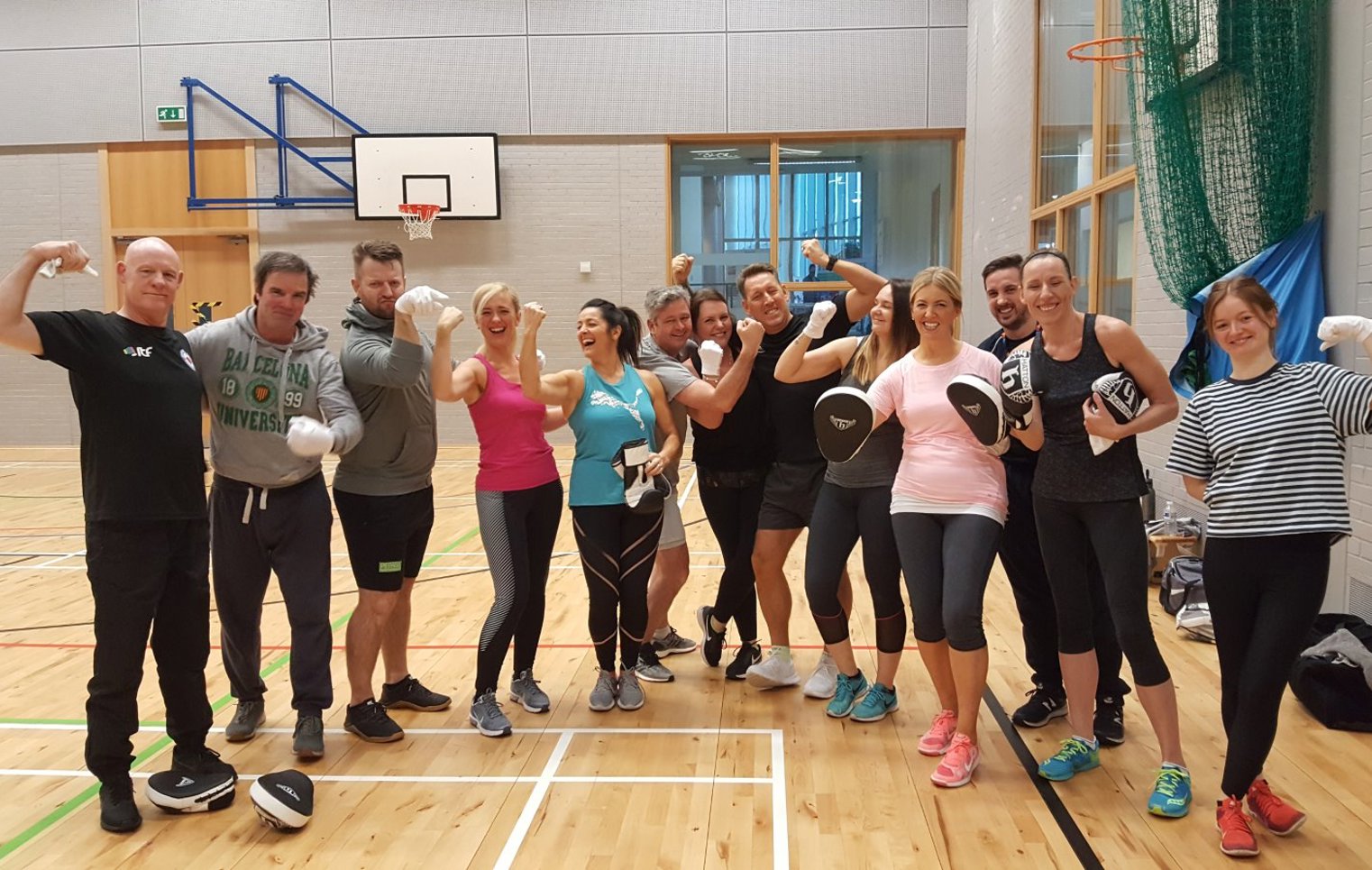 Group of Glasgow Kelvin College staff and students smiling and flexing after a fitness session in the gym hall, wearing boxing gloves and pads.