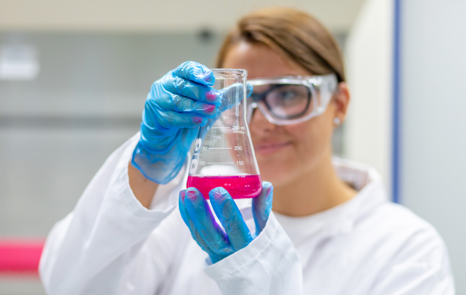 Student holding a beaker of pink liquid close to the camera, wearing gloves and safety goggles.