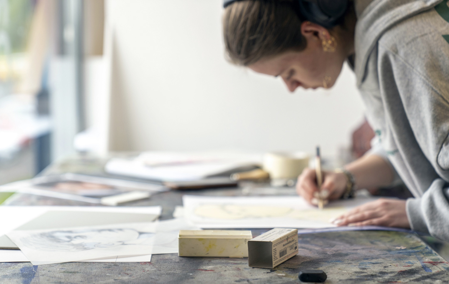 Person leaning over a table, drawing on paper in an art studio, with various sketches and art materials spread out