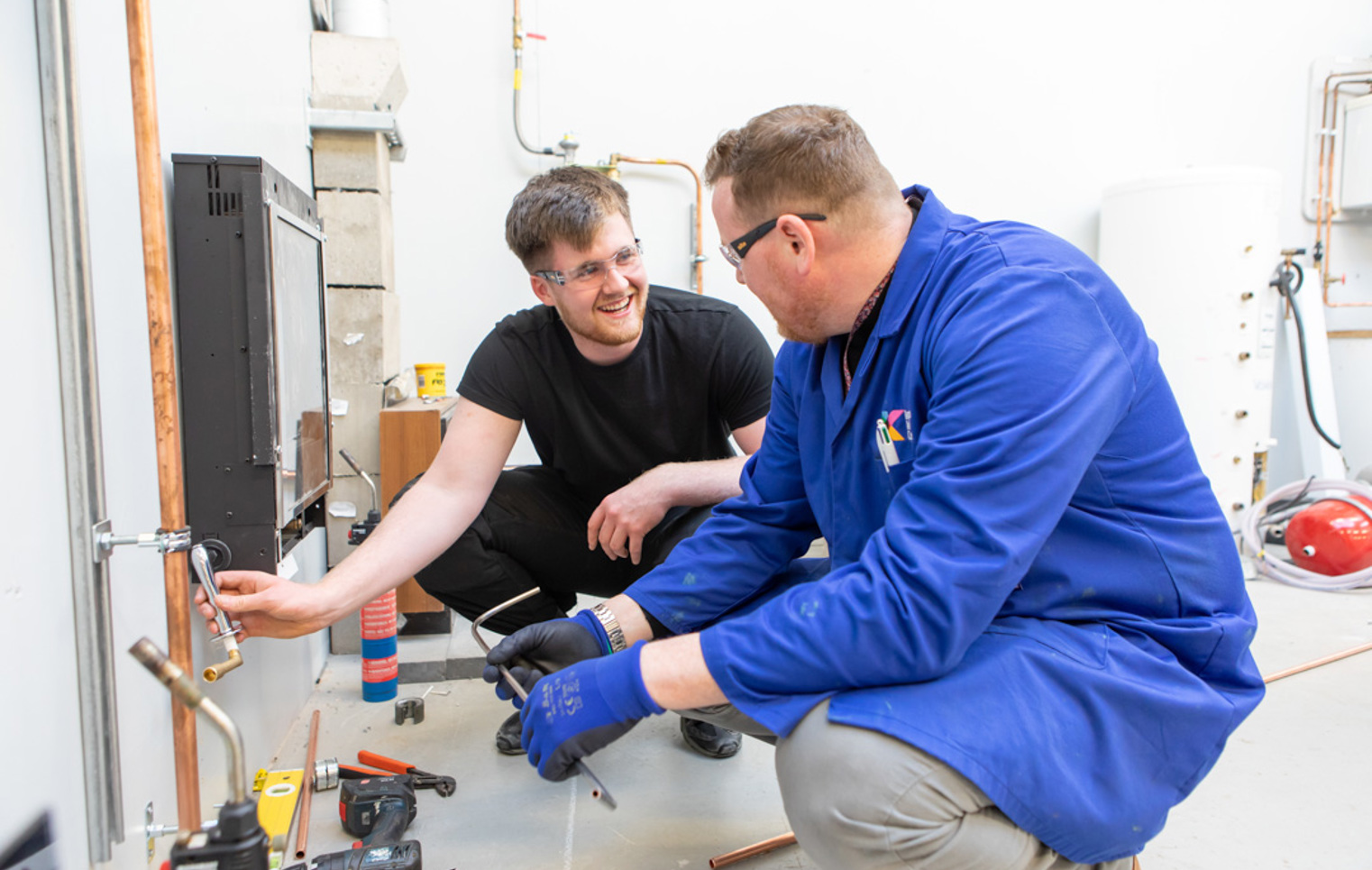 A plumbing lecturer and student smiling and collaborating closely while working together to install copper piping in a practical training session.