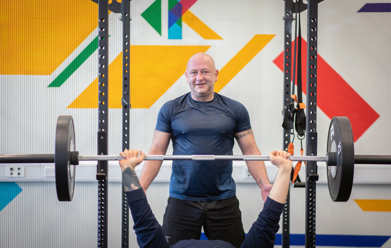 Student performing a bench press while another student spots them in a colourful training area.