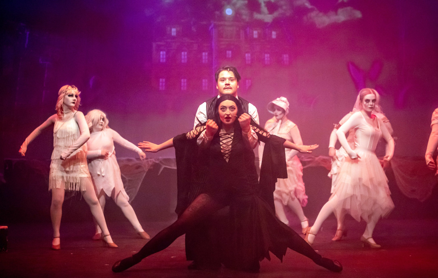 Group of musical theatre students performing a choreographed ensemble scene on stage, with central performers in dark costumes and dancers in white behind them.