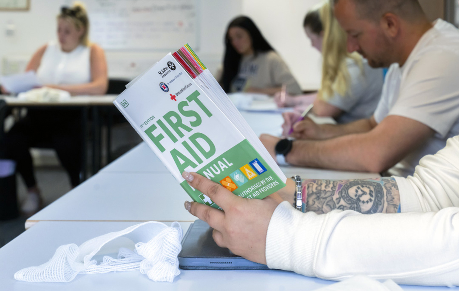 A close-up of a tattooed hand holding a first aid manual above a desk, with a white bandage and a tablet nearby—serious about learning, with a bit of style