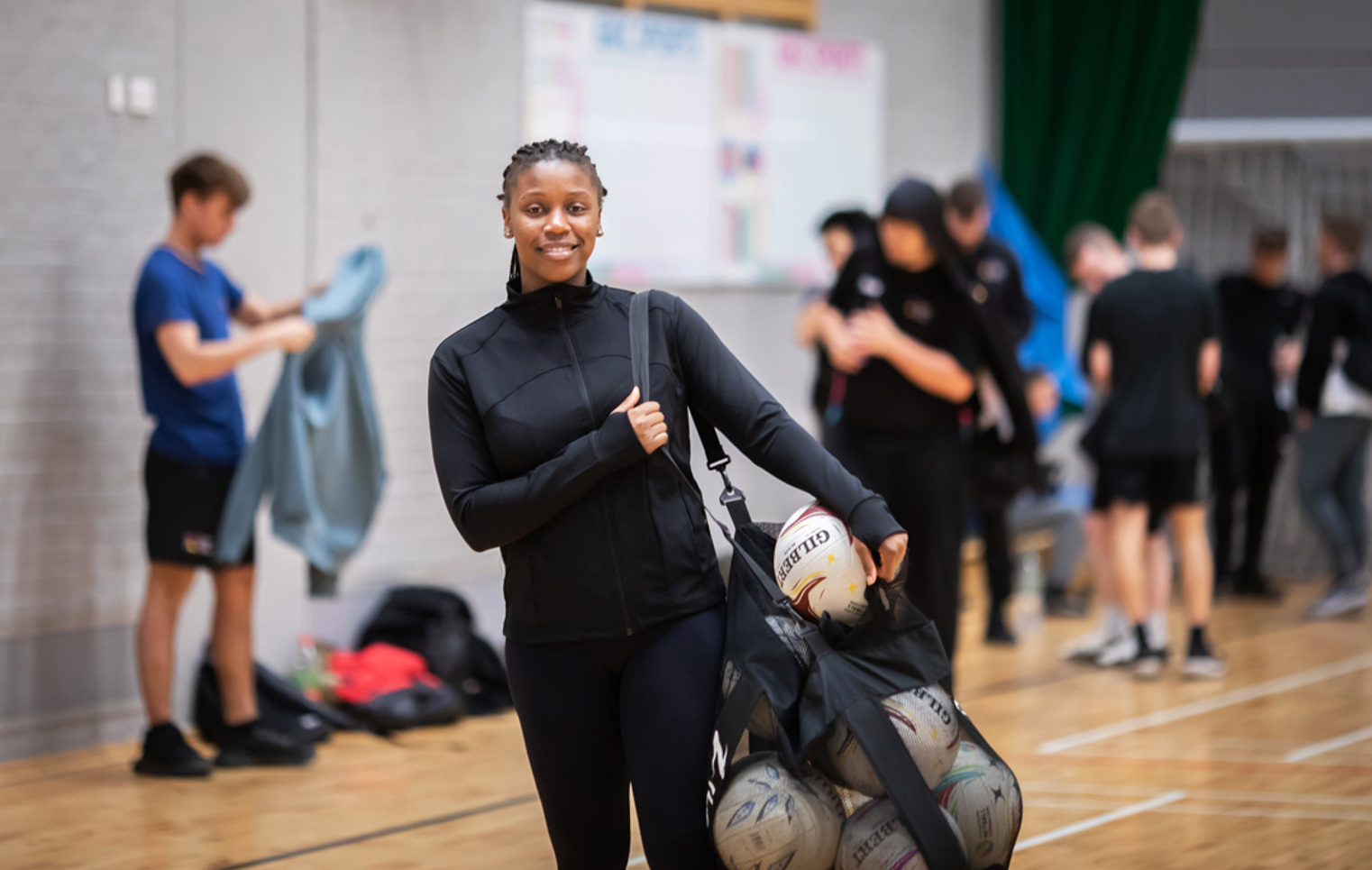 A student holding a mesh bag filled with netballs, smiling at the camera in a sports hall.