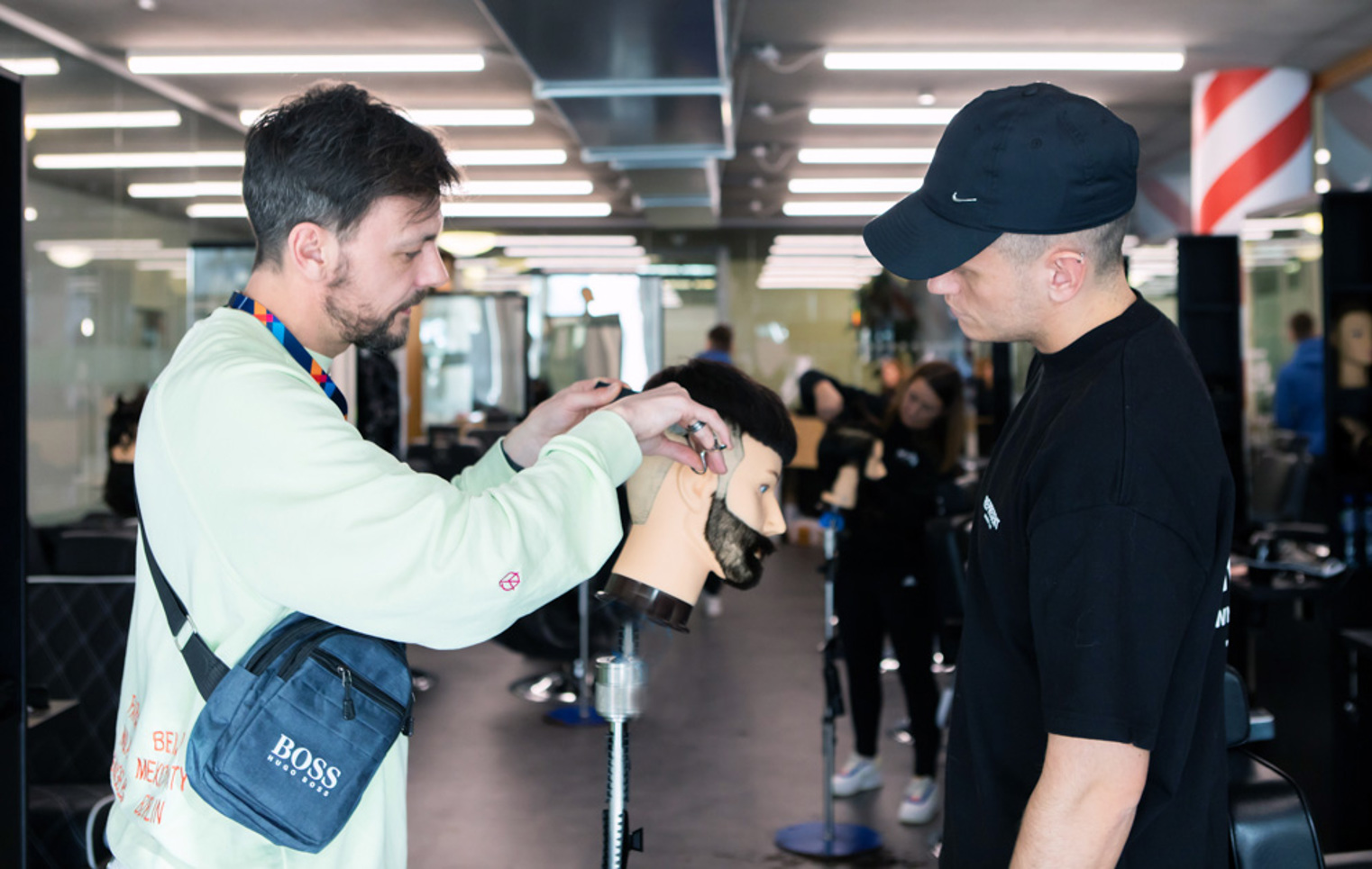 A barbering instructor demonstrating a haircut on a mannequin head while a student in a black cap observes.